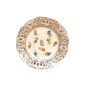Op de afbeelding: Een wit porseleinen bord met een gouden rand en een ingewikkeld bloemmotief. Het bord is versierd met kleine, kleurrijke bloemen.