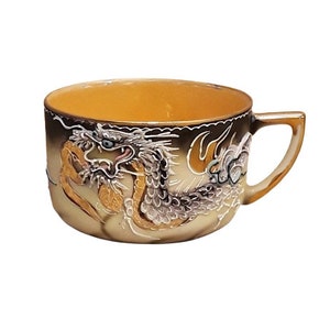 Può includere: Una tazza da tè in ceramica con un design di drago dorato e bianco. La tazza ha una forma arrotondata e un manico.