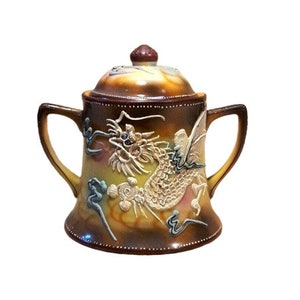 May include: A brown and yellow ceramic sugar bowl with a lid. The bowl is decorated with a detailed, raised image of a dragon with blue and white accents.
