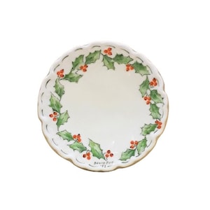 Puede incluir: Un plato de cerámica blanca con un borde festoneado y un diseño de hojas de acebo verdes y bayas rojas alrededor del borde. El plato está firmado "David Storr 1993" en la parte inferior.