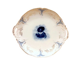 May include: A white porcelain serving platter with blue and gold accents. The platter features a central image of a person in a blue and white outfit wearing a hat. The platter has two handles on the sides.