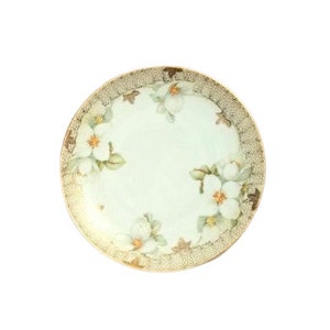 May include: A white porcelain plate with a gold rim and a floral design featuring white flowers with yellow centers. The flowers are arranged around the edge of the plate.