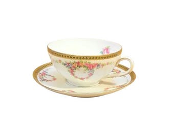 May include: A cream-coloured teacup and saucer set with gold trim and floral detailing. The teacup features a delicate handle and a central floral motif, while the saucer mirrors the gold rim and floral design. A classic set for tea.