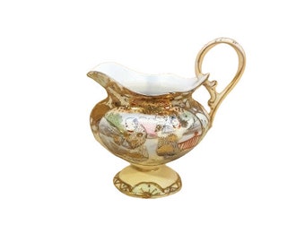 May include: A gold and white porcelain creamer with a delicate floral design and a handle. The creamer has a round body and a pedestal base.
