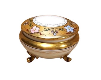 May include: A gold and white porcelain trinket box with a floral design and three gold feet. The lid has a white centre with a gold border and floral accents.