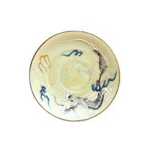 Op de afbeelding: Een witte keramische schotel met een gouden rand heeft een blauw en wit drakenontwerp. De draak is afgebeeld in een wervelende, dynamische pose met zijn staart gekruld rond het midden van de schotel.