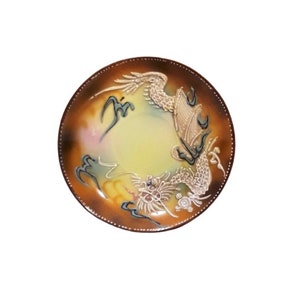 May include: A round ceramic plate with a brown, yellow, and white glaze. The plate features a detailed illustration of a dragon in flight.
