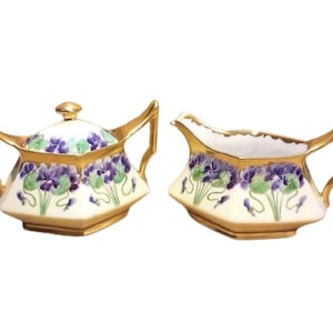 May include: A vintage porcelain sugar and creamer set with a delicate floral design. The set features a white background with purple and green violets and gold accents.