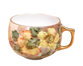 May include: A white porcelain teacup with a floral design. The cup has a rounded shape and a gold-trimmed handle. The floral design features yellow, orange, and green flowers on a light pink background.