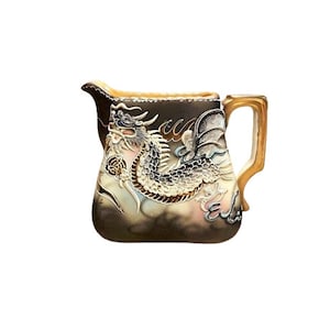 May include: A cream-colored ceramic creamer with a gold-colored handle. The creamer features a detailed embossed design of a dragon with gold accents.