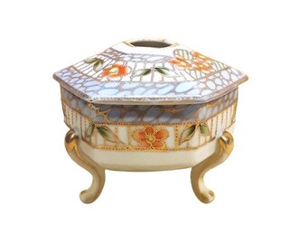 May include: A small, white porcelain trinket box with a hexagonal lid and three gold-painted feet. The lid is decorated with a floral pattern in shades of orange, green, and gold.