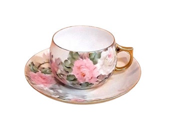 May include: A vintage teacup and saucer set with a floral design. The cup and saucer are white with a gold trim and feature pink roses and green leaves.