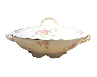 May include: A white porcelain serving dish with a lid, featuring a delicate floral pattern of pink roses and green leaves. The dish has two handles and a scalloped edge.