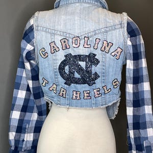 Op de afbeelding: Een korte spijkerjas met blauw-wit geruite mouwen. De achterkant van de jas toont de woorden "CAROLINA TAR HEELS" in roze en blauwe paillettenletters, met een bijpassend logo in het midden.