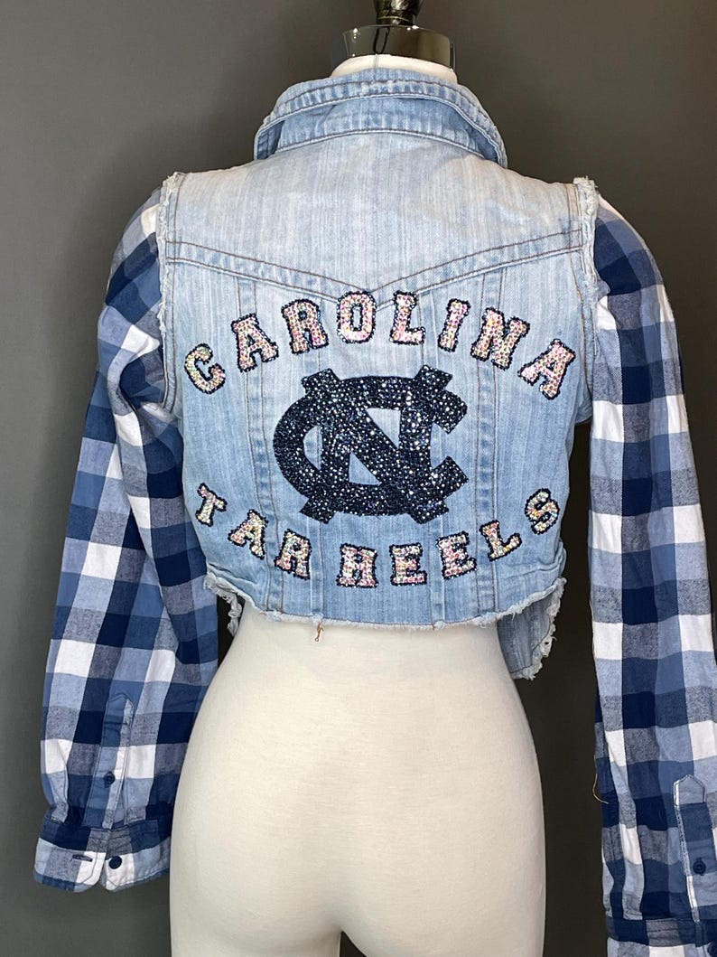 Op de afbeelding: Een korte spijkerjas met blauw-wit geruite mouwen. De achterkant van de jas toont de woorden "CAROLINA TAR HEELS" in roze en blauwe paillettenletters, met een bijpassend logo in het midden.