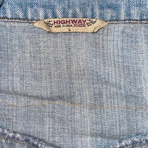 Op de afbeelding: Close-up van een lichtblauwe spijkerjas. Het label leest "HIGHWAY MADE IN CHINA Jeans" met maat "L". De denim stof heeft zichtbare stiksels en een getextureerd uiterlijk, typerend voor spijkerjassen.