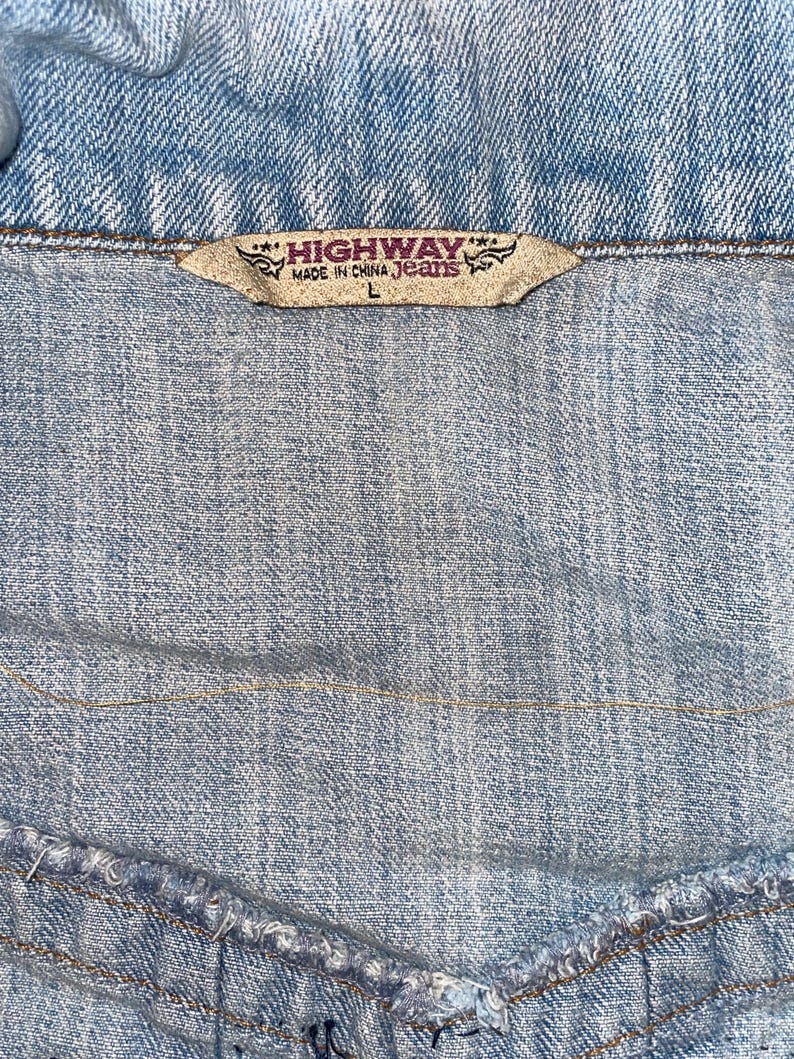 Op de afbeelding: Close-up van een lichtblauwe spijkerjas. Het label leest "HIGHWAY MADE IN CHINA Jeans" met maat "L". De denim stof heeft zichtbare stiksels en een getextureerd uiterlijk, typerend voor spijkerjassen.
