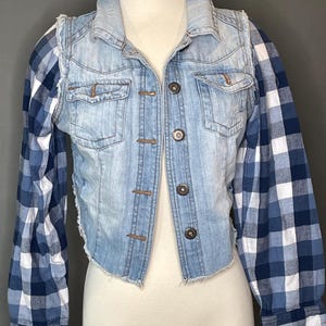 Op de afbeelding: Een lichtblauwe spijkerbroekvest met een gerafelde zoom en knopen. Het vest heeft lange mouwen van een blauw-wit geruite stof. De jas wordt op een mannequin getoond.