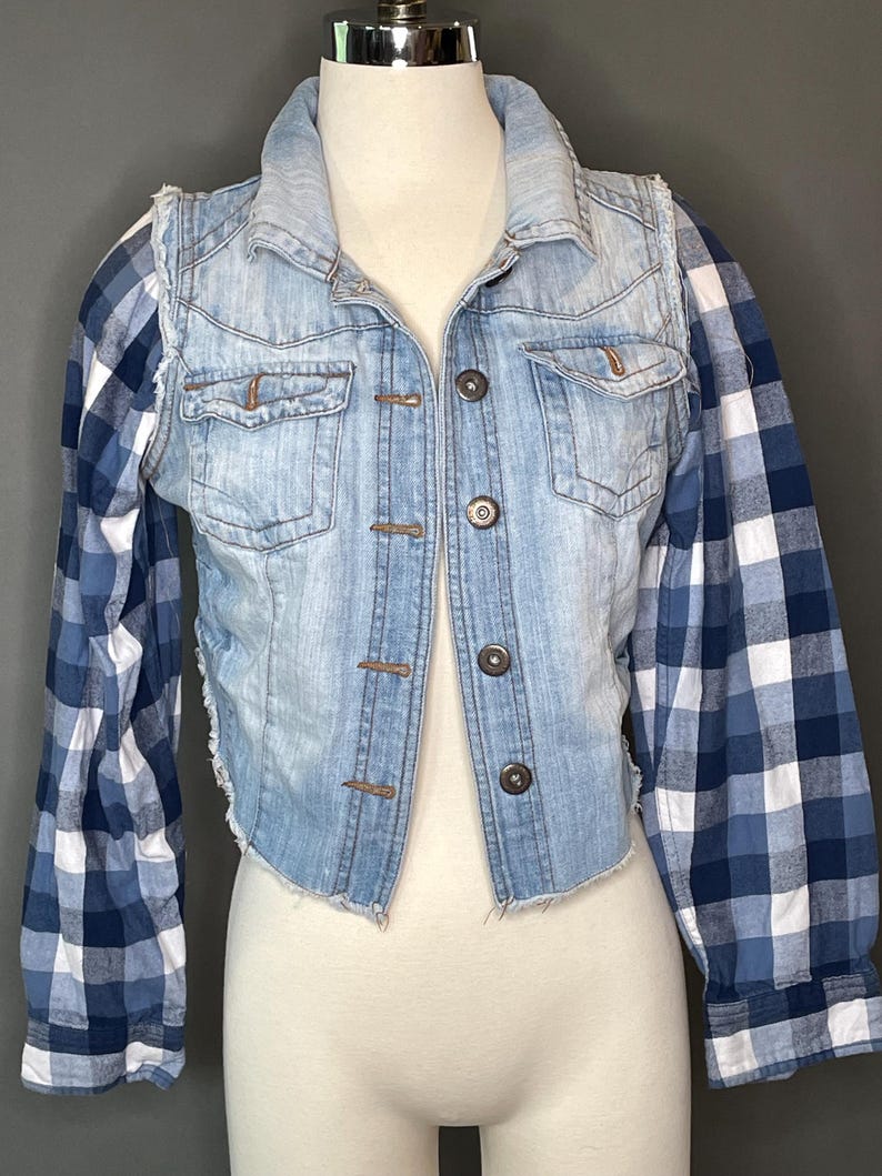 Op de afbeelding: Een lichtblauwe spijkerbroekvest met een gerafelde zoom en knopen. Het vest heeft lange mouwen van een blauw-wit geruite stof. De jas wordt op een mannequin getoond.