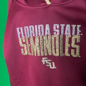 以下が含まれることがあります： キラキラ光る銀と金の文字で「FLORIDA STATE SEMINOLES」と書かれたマルーン色のパーカー。 FSUのロゴがテキストの下にあります。 フードには金のトリムが付いています。