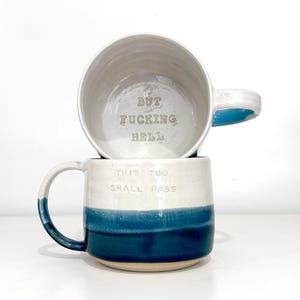 May include: Two ceramic mugs stacked on a white surface. The bottom mug is white with a blue band and the text "THIS TOO SHALL PASS." The top mug is upside down, revealing the text "BUT FUCKING HELL."