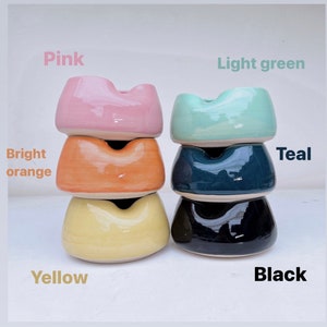 May include: Stack of four ceramic ashtrays in different colors: pink, bright orange, yellow, and black. The top ashtray is light green and the second from the top is teal.