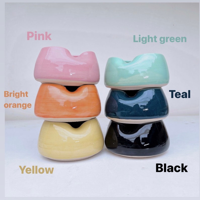 May include: Stack of four ceramic ashtrays in different colors: pink, bright orange, yellow, and black. The top ashtray is light green and the second from the top is teal.