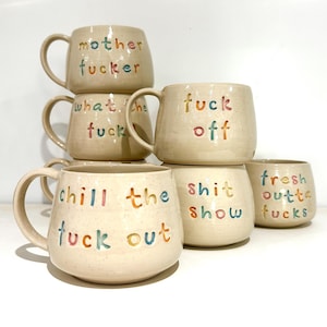 May include: A collection of six speckled cream-coloured ceramic mugs, each featuring colourful, embossed lettering. The mugs display various phrases including mother fucker, what the fuck, fuck off, chill the fuck out, shit show, and fresh outta fucks.