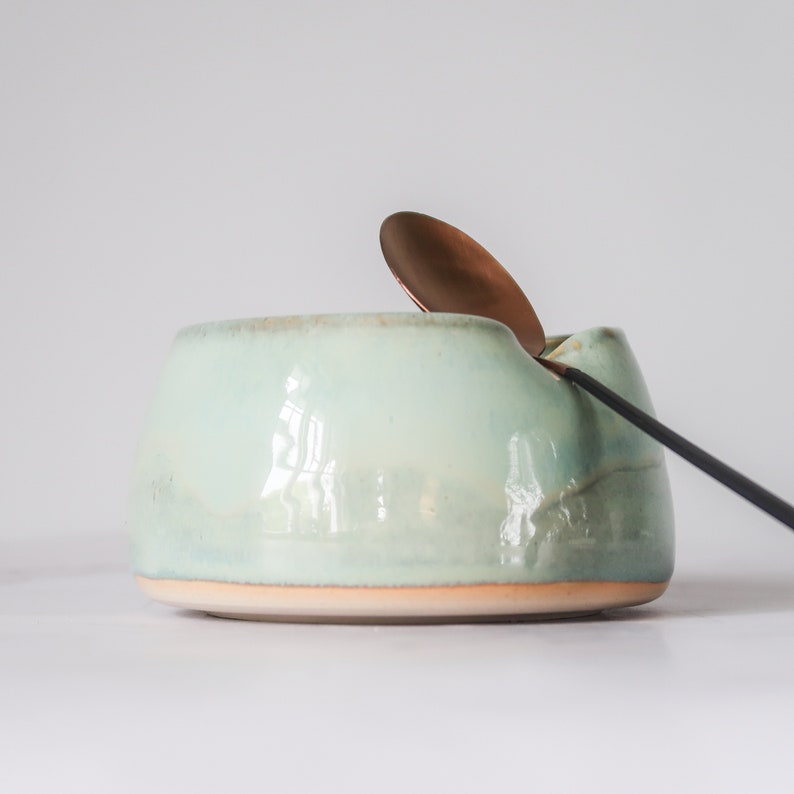 May include: A light blue ceramic bowl with a rounded shape and a slightly uneven rim. A copper spoon rests on the edge of the bowl.