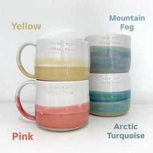 May include: Four ceramic mugs stacked, each with a different color scheme. The mugs are white with a colored band at the bottom and the text "THIS TOO SHALL PASS". Colors include yellow, pink, mountain fog, and arctic turquoise.