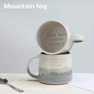 May include: Two ceramic mugs stacked on top of each other. The bottom mug has a gradient design in shades of gray, green, and white. The text "HAPPY FIRST HOME AMY & SAM 19.05.2021" is visible. The top mug has the text "LOVE YOUS LOADS, NIMMS X". The text "Mountain fog" is at the top.