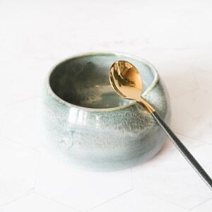 May include: A light blue ceramic bowl with a gold spoon. The bowl has a textured surface and the spoon has a black handle.