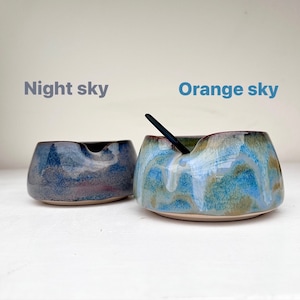 May include: Two ceramic bowls with a blue and green glaze. The bowl on the right has a black spoon inside. The bowl on the left has a darker blue glaze. The text "Night sky" is above the left bowl and the text "Orange sky" is above the right bowl.
