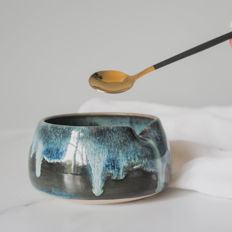 May include: A dark blue and green ceramic bowl with a white glaze. The bowl has a unique shape with a dip on one side. A gold spoon is hovering above the bowl.