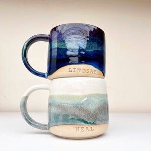 May include: Two ceramic mugs with a blue and green glaze. The top mug has the name "LINDSAY" on it and the bottom mug has the name "NEAL" on it.