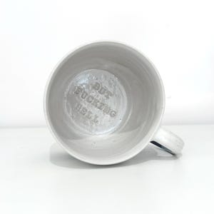 May include: A white ceramic mug with a handle, viewed from above. The interior of the mug has the words "BUT FUCKING HELL" embossed in the bottom. The mug is set against a white background.