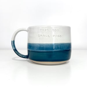 May include: A ceramic mug with a white upper half and a deep blue lower half. The mug has a curved handle and the words "THIS TOO SHALL PASS" are embossed on the front.