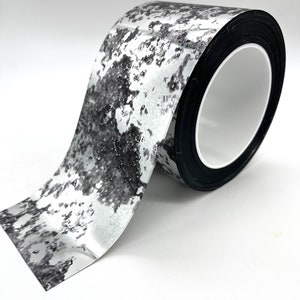 Puede incluir: Un rollo de cinta adhesiva con estampado plateado y negro con un núcleo negro.