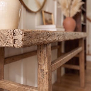 May include: A rustic, weathered wooden console table with a natural finish. The table features a rectangular top and four sturdy legs. A cream-colored lamp and a terracotta vase with dried plants sit on the table, adding to the decor.