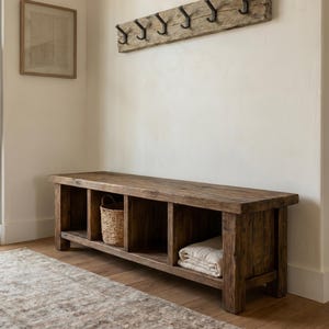 May include: A long, rectangular wooden bench with four open storage compartments, featuring a woven basket and a folded white textile inside. Above the bench, a horizontal piece of reclaimed wood is mounted to the wall with six black metal hooks.
