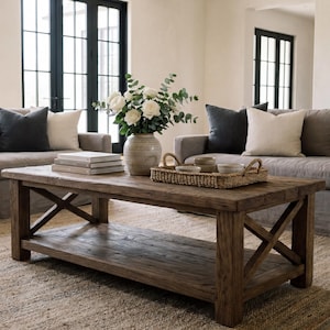 May include: A living room scene featuring two gray sofas with dark and light pillows, a rustic wooden coffee table, and a woven rug. A vase of white flowers and greenery sits on the table, with a woven tray holding small bowls.