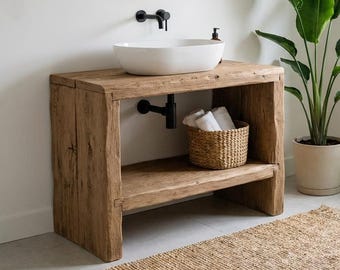 May include: A bathroom vanity made of light brown reclaimed wood with a white oval vessel sink and a matte black faucet. The vanity features an open shelf holding a woven basket with white towels. A round wooden mirror hangs above the sink.