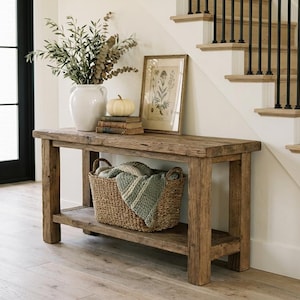 May include: A rustic wooden console table with a lower shelf, holding a woven basket with a blanket. A white vase with greenery, a small pumpkin, and framed artwork sit on top. The scene is next to a staircase.