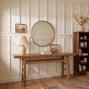 May include: A neutral-toned entryway featuring a wooden console table, a round gold mirror, and a cream-colored lamp with a pleated shade. A dark wood bookshelf is on the right, and framed artwork hangs on the wall. The room has a rug and a staircase.