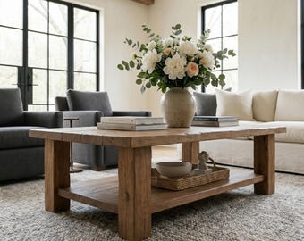 May include: A rectangular wooden coffee table with a lower shelf, featuring a light brown finish and a thick, sturdy frame. On the tabletop sits a ceramic vase filled with white and peach flowers, alongside two stacked books and a small bowl.
