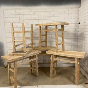 May include: Several small, light-colored wooden stools are arranged in a room. The stools have a simple design with rectangular seats and four angled legs. The wood grain is visible, and the stools appear to be handmade.
