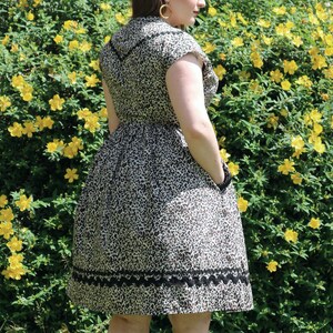 Puede incluir: Una mujer lleva un vestido de estampado de leopardo marr&oacute;n y negro con ribete de encaje negro. El vestido tiene manga corta y cuello. Est&aacute; de pie frente a un arbusto de flores amarillas.