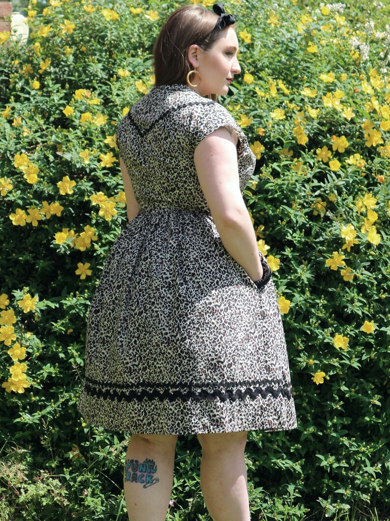 Puede incluir: Una mujer lleva un vestido de estampado de leopardo marr&oacute;n y negro con ribete de encaje negro. El vestido tiene manga corta y cuello. Est&aacute; de pie frente a un arbusto de flores amarillas.