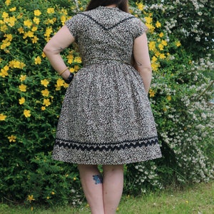 Puede incluir: Una mujer lleva un vestido de estampado de leopardo marr&oacute;n y negro con un ribete de encaje negro. El vestido tiene manga corta y un corpi&ntilde;o ajustado. La mujer est&aacute; de pie frente a un arbusto verde con flores amarillas.