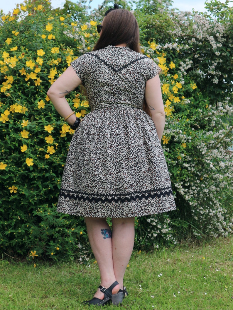 Puede incluir: Una mujer lleva un vestido de estampado de leopardo marr&oacute;n y negro con un ribete de encaje negro. El vestido tiene manga corta y un corpi&ntilde;o ajustado. La mujer est&aacute; de pie frente a un arbusto verde con flores amarillas.